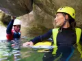 Kobieta w żółtym kasku i piankowym stroju zanurzona w wodzie, obok mężczyzna w białym kasku. To właśnie canyoning – przygoda w kanionie.