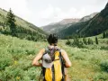 Kobieta z plecakiem wyrusza na trekking przez kwiecistą dolinę, podziwiając górskie krajobrazy.