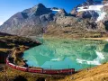 Czerwony pociąg wije się wzdłuż turkusowego jeziora u podnóża alpejskich szczytów. Idealne miejsce na trekking w alpach.
