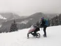 Zimowe Tatry z wózkiem. Kobieta w czapce i z plecakiem pcha wózek po ośnieżonym stoku, podziwiając górski krajobraz.