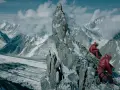 Dwóch alpinistów w czerwonych kurtkach odpoczywa na skalistym szczycie, podziwiając lodowcową dolinę i ośnieżone góry.