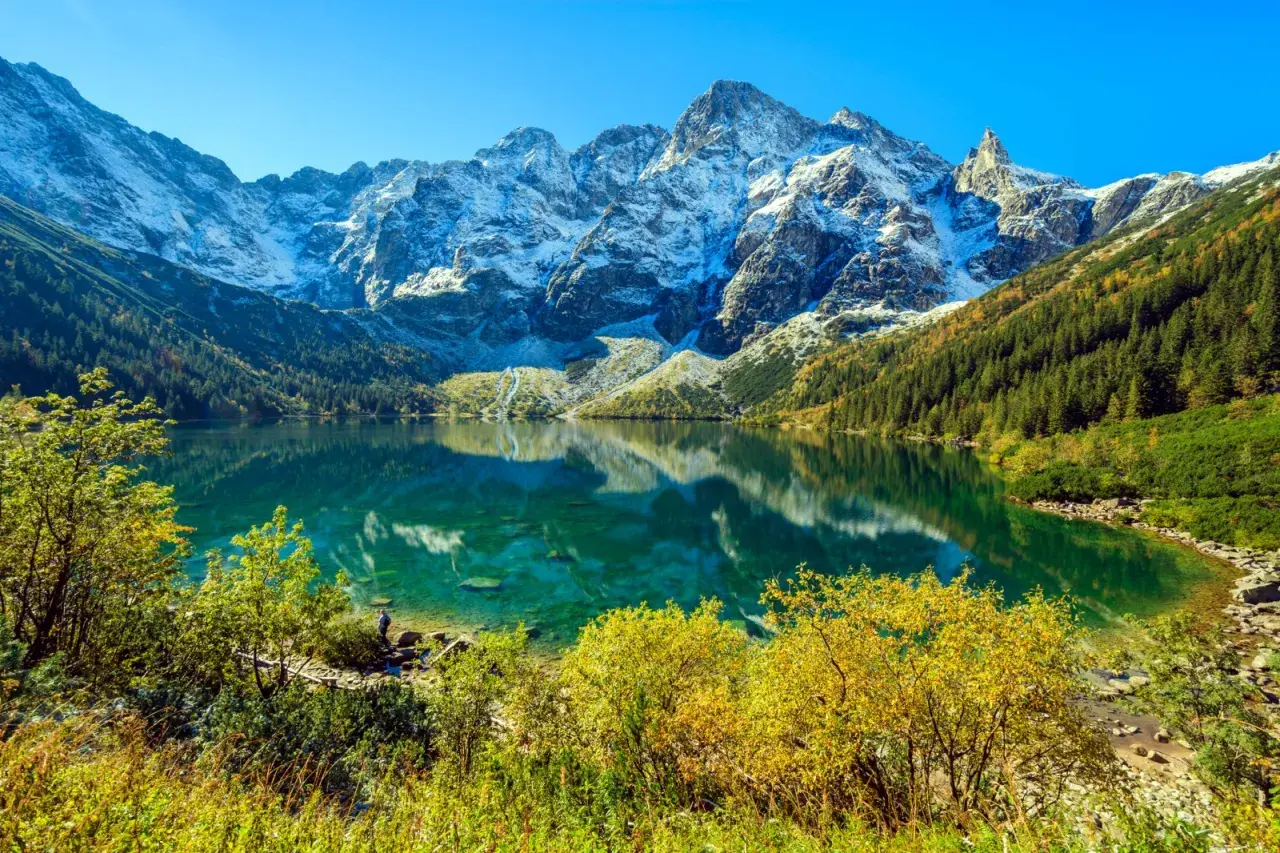 Turkusowe Morskie Oko odbija ośnieżone szczyty Tatr. Jesienne drzewa otaczają taflę wody.