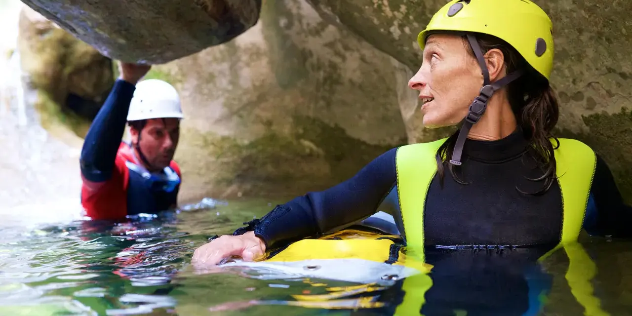 Kobieta w żółtym kasku i piankowym stroju zanurzona w wodzie, obok mężczyzna w białym kasku. To właśnie canyoning – przygoda w kanionie.