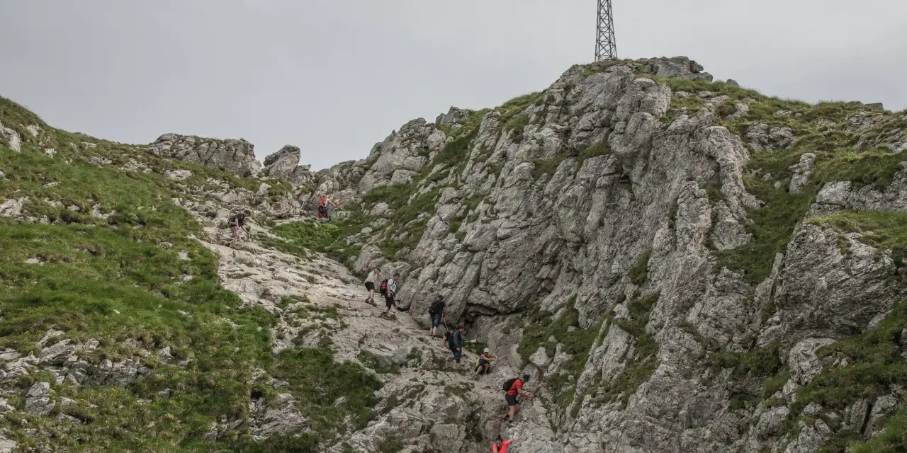 Grupa turystów wspina się po skalistym zboczu w kierunku krzyża na szczycie. Trudno określić, ile idzie się na Giewont, ale widok jest imponujący.