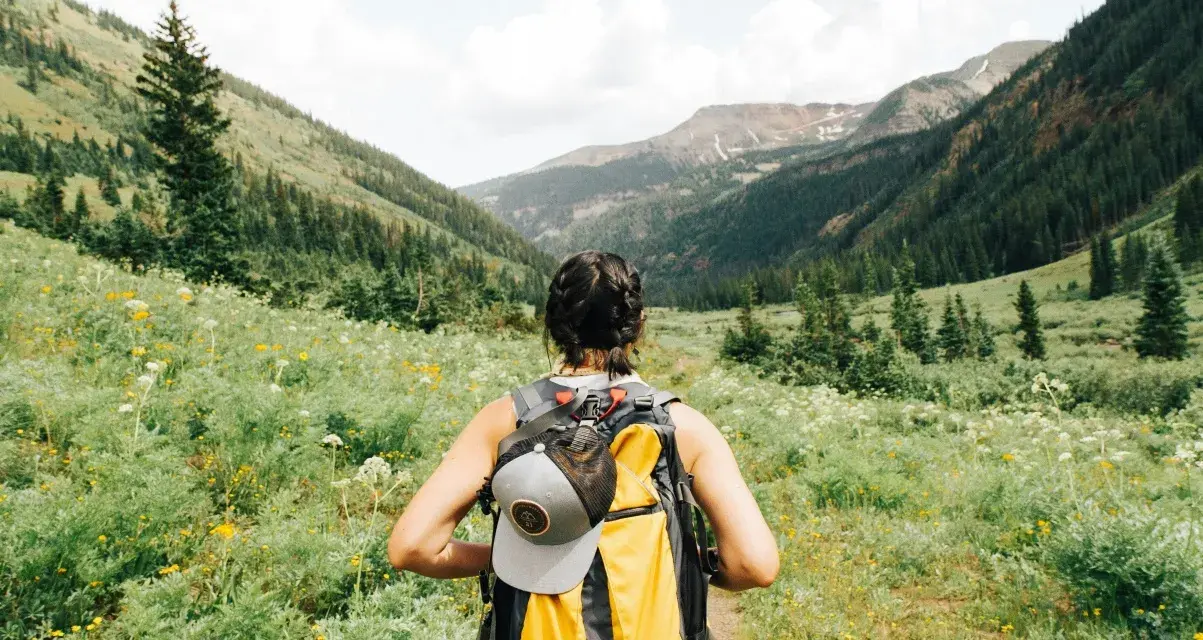 Kobieta z plecakiem wyrusza na trekking przez kwiecistą dolinę, podziwiając górskie krajobrazy.
