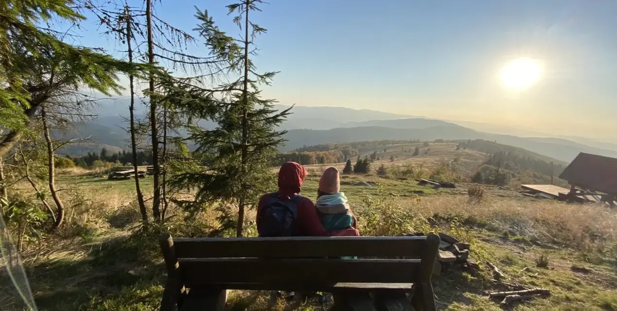 Para siedzi na ławce, podziwiając widok na góry. Słońce zachodzi nad horyzontem, malując niebo na złote i pomarańczowe barwy. To piękny widok z mogielicy z Jurkowa.