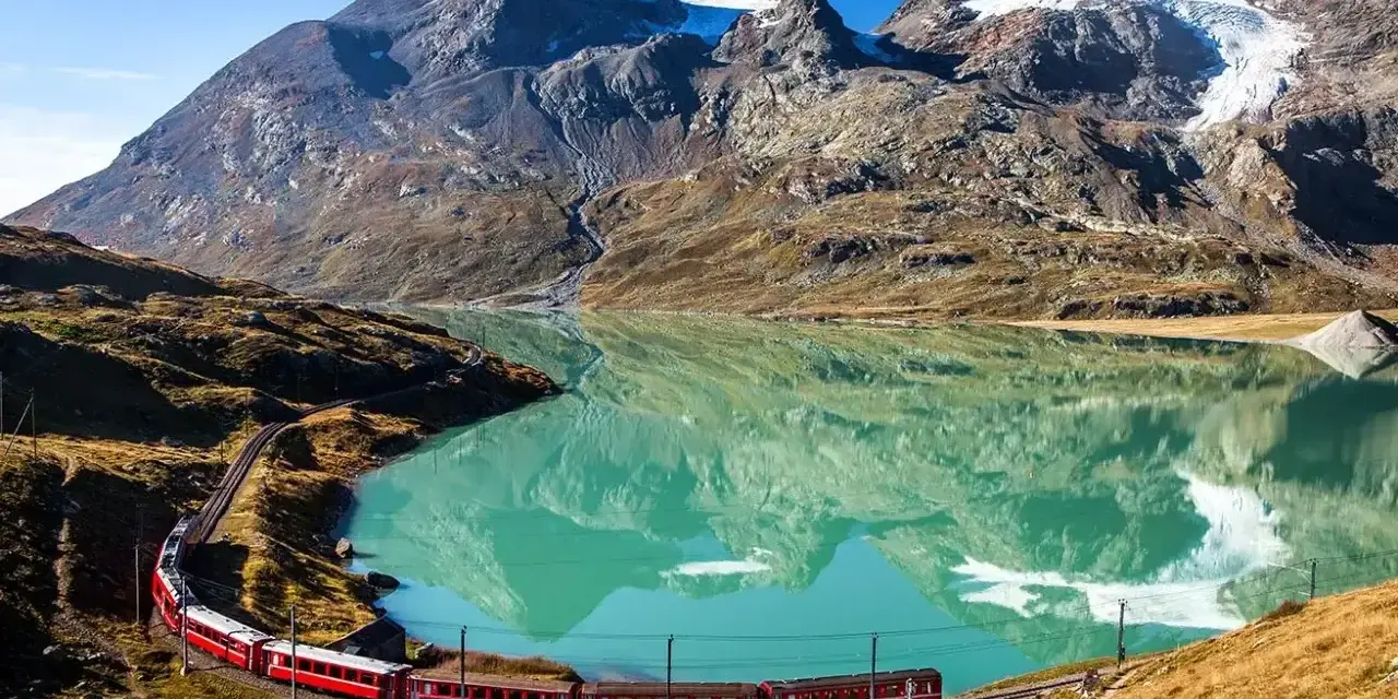 Czerwony pociąg wije się wzdłuż turkusowego jeziora u podnóża alpejskich szczytów. Idealne miejsce na trekking w alpach.