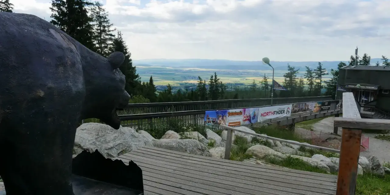 Pomnik niedźwiedzia na tle panoramy Tatr poza szlakiem. Drewniana kładka prowadzi do budynku z widokiem na dolinę.