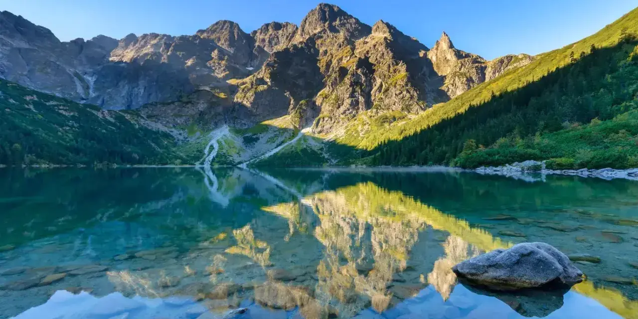 Idealny weekend w Tatrach: Morskie Oko odbija skaliste szczyty i zielone zbocza. Czysta woda odsłania kamienie.