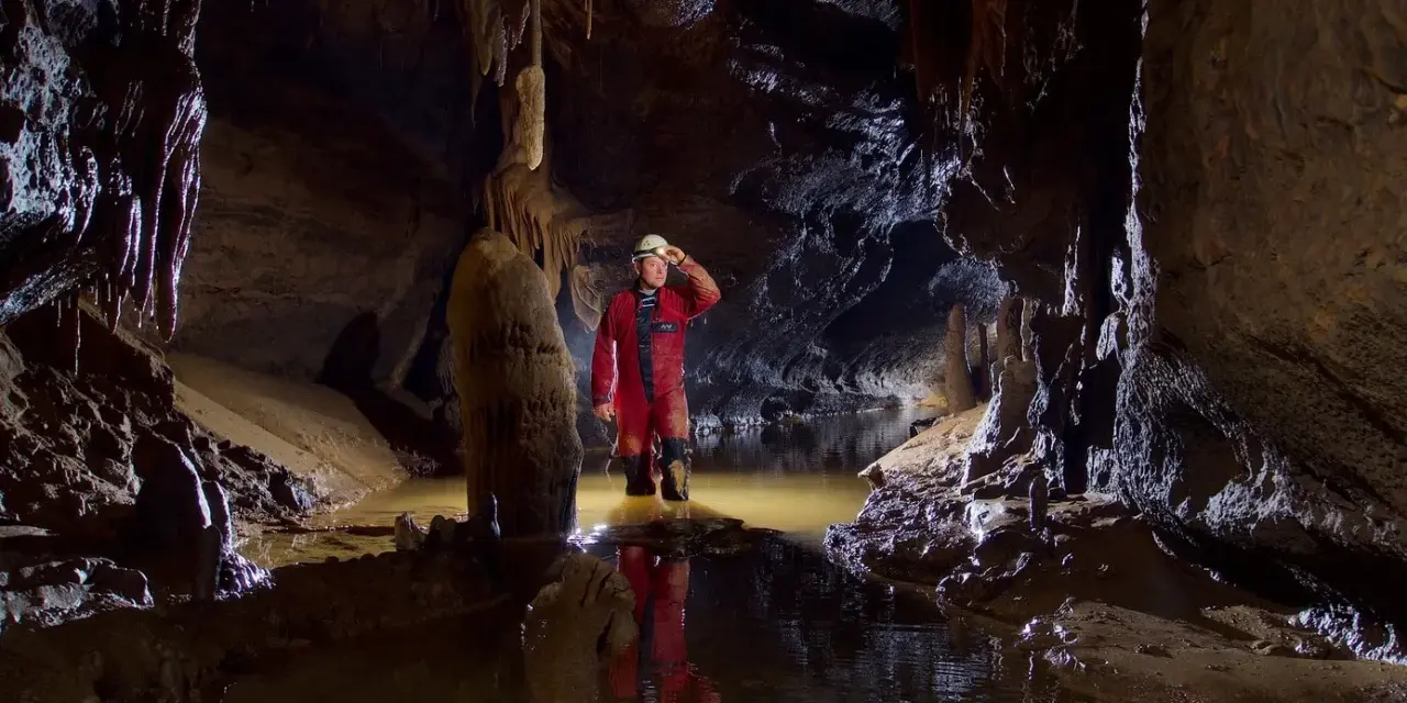 Eksplorator w jaskini, zanurzony po kolana w wodzie, odkrywa tajemnice speleologii. Stalagmity i nacieki tworzą niezwykły krajobraz.