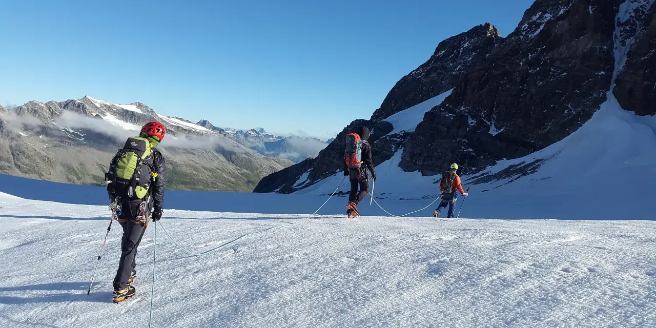 Trzej alpiniści na lodowcu, uczący się jak zostać przewodnikiem górskim. W tle majestatyczne szczyty.