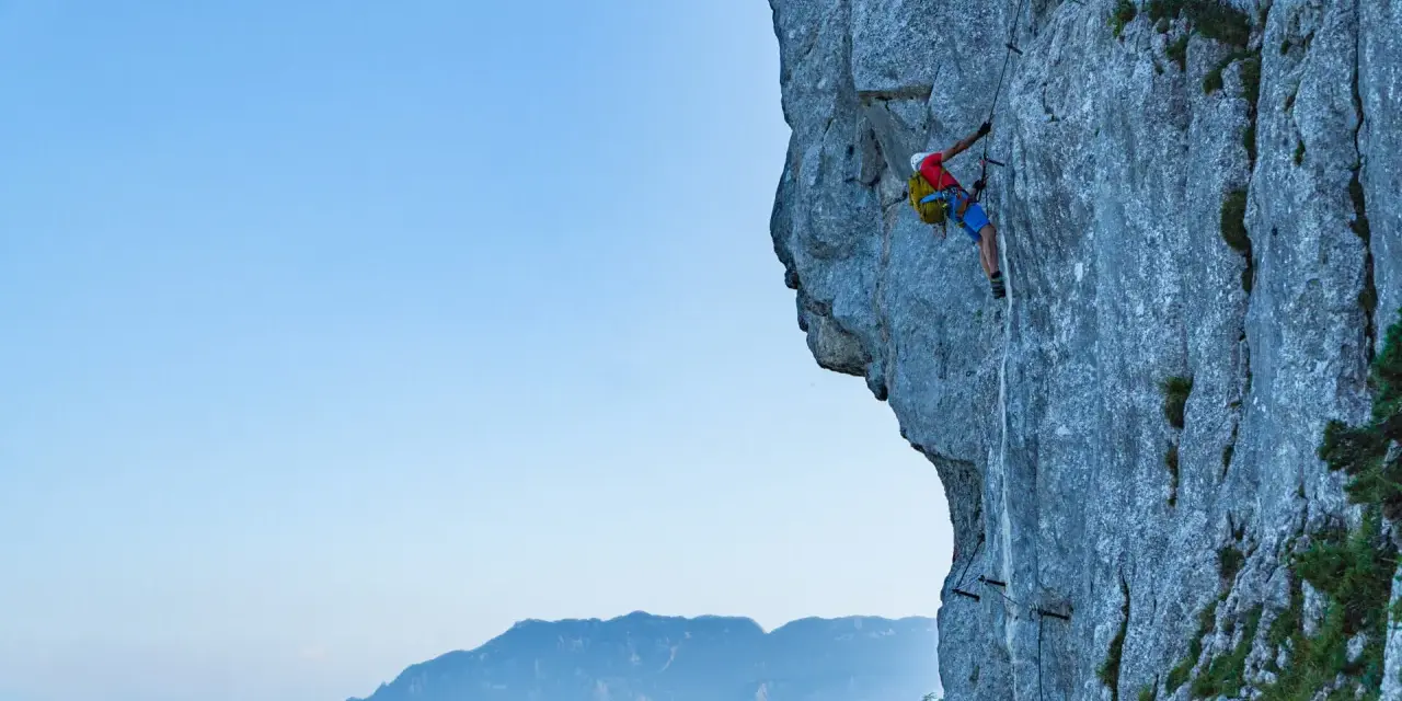 Wspinacz pokonuje via ferrata niemcy, zmagając się z pionową ścianą skalną. W tle piękne góry.