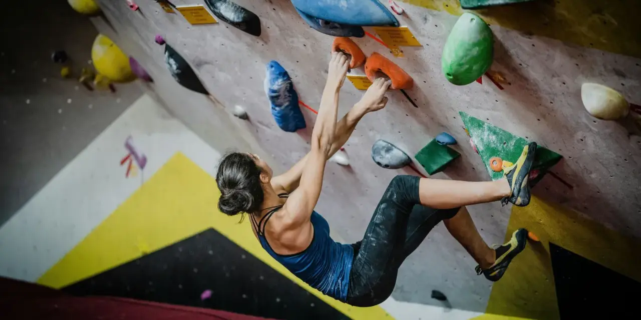 Kobieta wspina się po kolorowej ścianie, szukając chwytów. To właśnie bouldering – sportowa wspinaczka bez liny, na niskich ścianach.