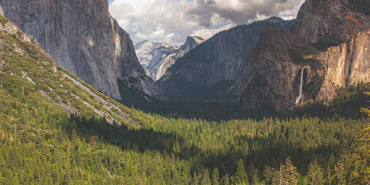 Majestatyczne klify i wodospad w **parku narodowym Yosemite**, otoczone gęstym lasem iglastym.