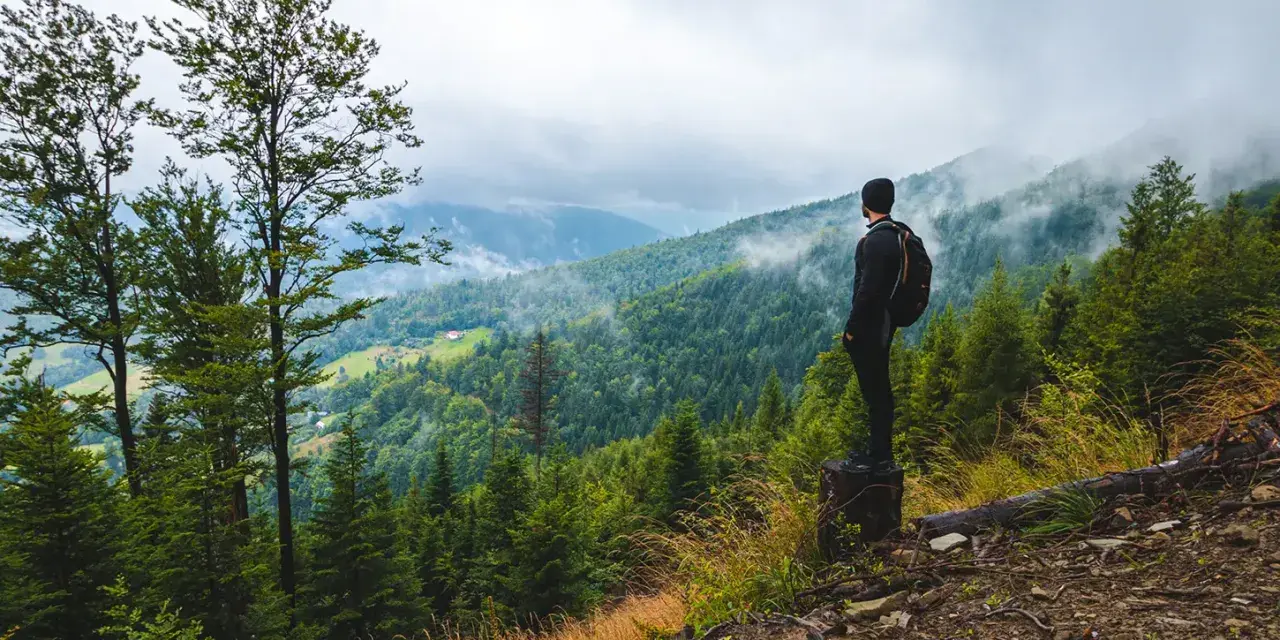 Mężczyzna z plecakiem podziwia mglisty górski krajobraz. To właśnie jest trekking – odkrywanie natury pieszo.