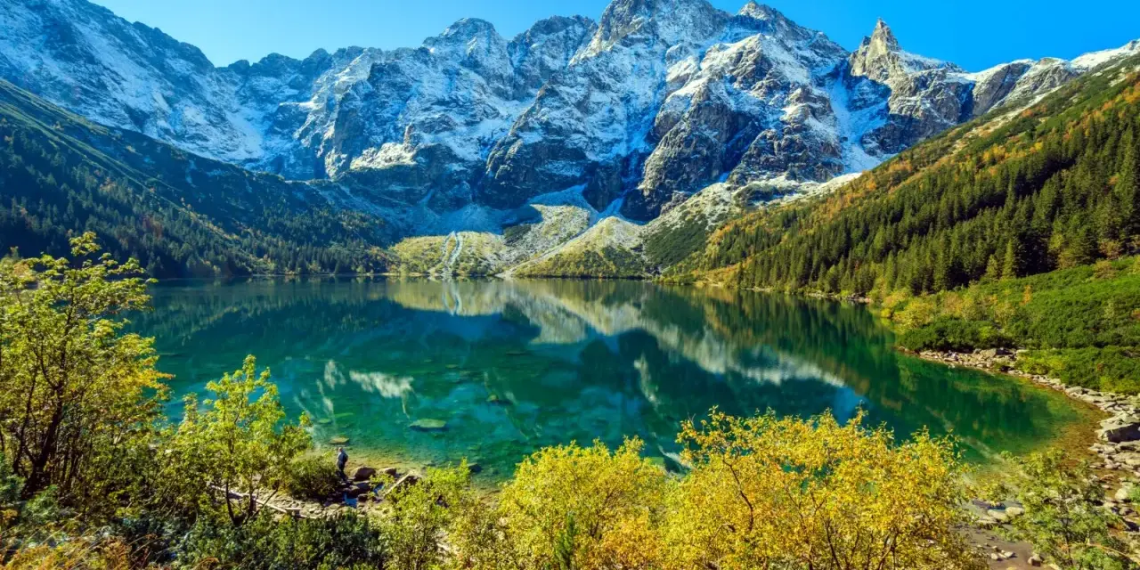 Turkusowe Morskie Oko odbija ośnieżone szczyty Tatr. Jesienne drzewa otaczają taflę wody.