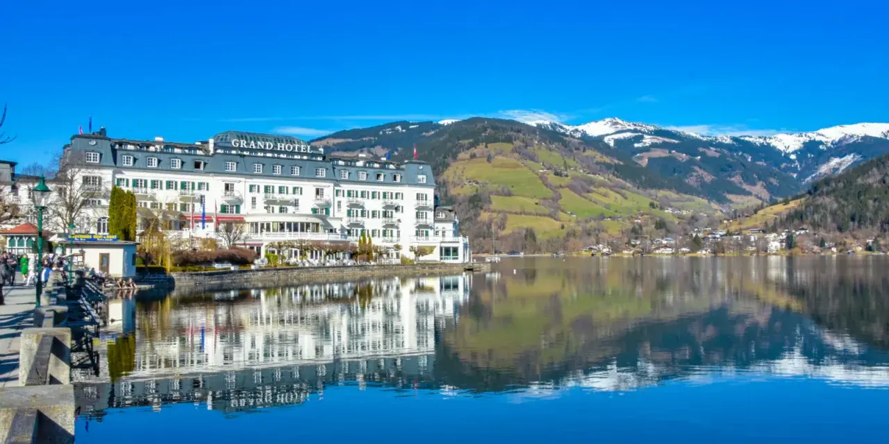 Grand Hotel nad jeziorem w Zell am See, z odbiciem w spokojnej wodzie i ośnieżonymi górami w tle.