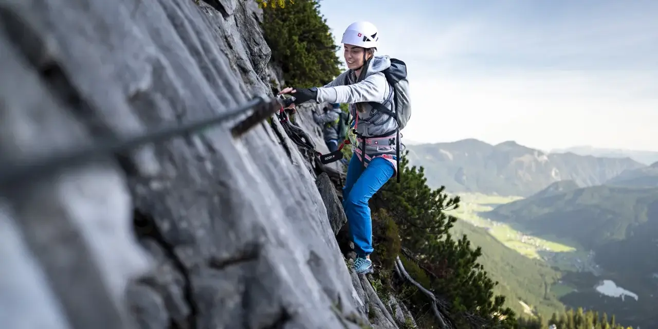 Kobieta w kasku i uprzęży pokonuje skalną ścianę, korzystając z liny. To właśnie jest via ferrata – bezpieczna droga wspinaczkowa.