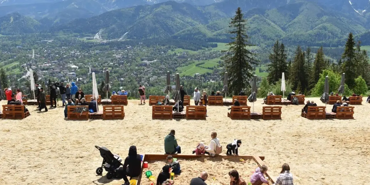 Piękny widok z Gubałówki, gdzie turyści odpoczywają na piasku. Ile metrów do szczytu? Tego nie wiemy, ale widok jest wart każdej wysokości.