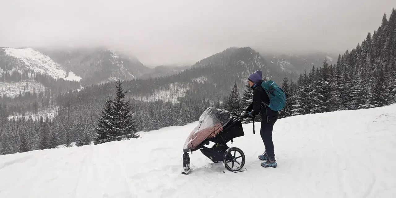 Zimowe Tatry z wózkiem. Kobieta w czapce i z plecakiem pcha wózek po ośnieżonym stoku, podziwiając górski krajobraz.