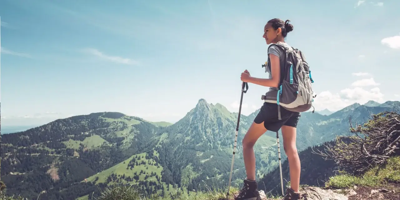 Młoda kobieta z plecakiem i kijkami trekkingowymi podziwia górski krajobraz. Pokazuje, jak chodzić z kijkami trekkingowymi, by czerpać radość z wędrówki.