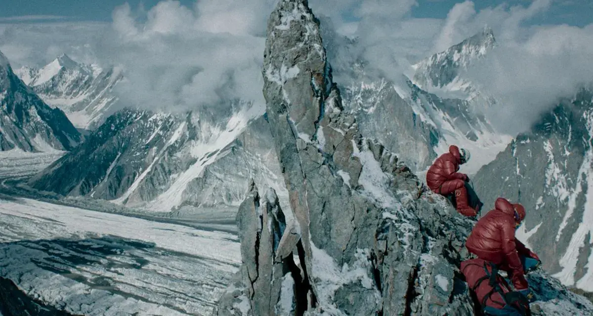 Dwóch alpinistów w czerwonych kurtkach odpoczywa na skalistym szczycie, podziwiając lodowcową dolinę i ośnieżone góry.