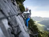 Kobieta w kasku i uprzęży pokonuje skalną ścianę, korzystając z liny. To właśnie jest via ferrata – bezpieczna droga wspinaczkowa.