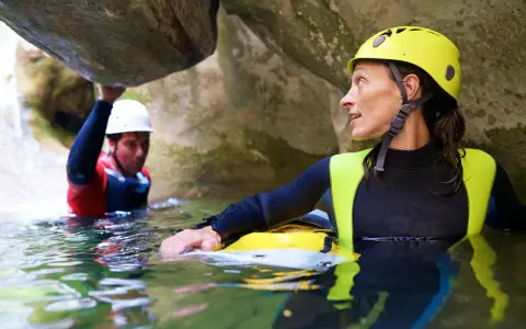 Kobieta w żółtym kasku i piankowym stroju zanurzona w wodzie, obok mężczyzna w białym kasku. To właśnie canyoning – przygoda w kanionie.