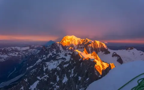 Mont Blanc latem, oświetlony złotym blaskiem wschodu słońca, z cieniem góry rzucającym się na niebo.