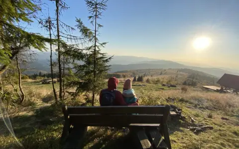 Para siedzi na ławce, podziwiając widok na góry. Słońce zachodzi nad horyzontem, malując niebo na złote i pomarańczowe barwy. To piękny widok z mogielicy z Jurkowa.