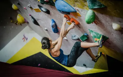 Kobieta wspina się po kolorowej ścianie, szukając chwytów. To właśnie bouldering – sportowa wspinaczka bez liny, na niskich ścianach.