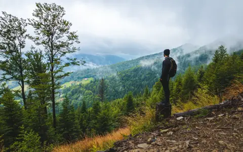 Mężczyzna z plecakiem podziwia mglisty górski krajobraz. To właśnie jest trekking – odkrywanie natury pieszo.