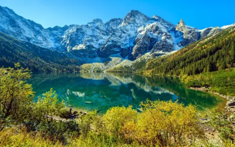 Turkusowe Morskie Oko odbija ośnieżone szczyty Tatr. Jesienne drzewa otaczają taflę wody.