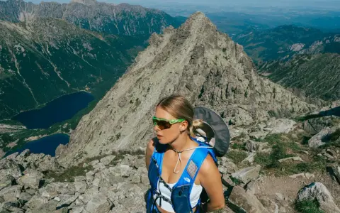 Wyprawa w góry zakończona sukcesem. Młoda kobieta w okularach przeciwsłonecznych i niebieskim plecaku podziwia malowniczy widok na jeziora i skaliste szczyty.