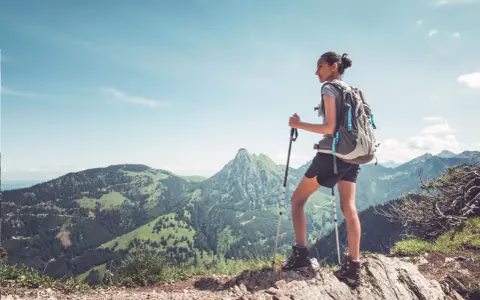 Młoda kobieta z plecakiem i kijkami trekkingowymi podziwia górski krajobraz. Pokazuje, jak chodzić z kijkami trekkingowymi, by czerpać radość z wędrówki.