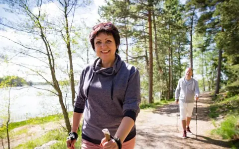 Uśmiechnięta kobieta i mężczyzna spacerują z kijkami po leśnej ścieżce. Nie ma widocznych przeciwwskazań do chodzenia z kijkami.