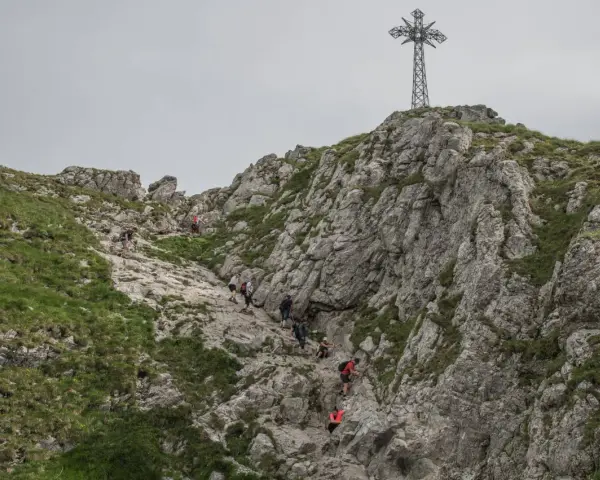 Grupa turystów wspina się po skalistym zboczu w kierunku krzyża na szczycie. Trudno określić, ile idzie się na Giewont, ale widok jest imponujący.