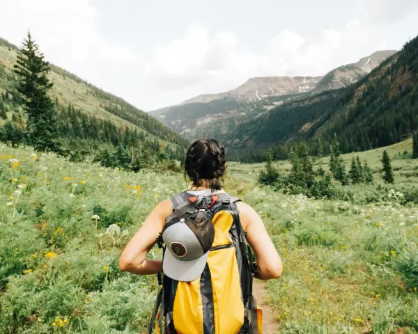 Kobieta z plecakiem wyrusza na trekking przez kwiecistą dolinę, podziwiając górskie krajobrazy.