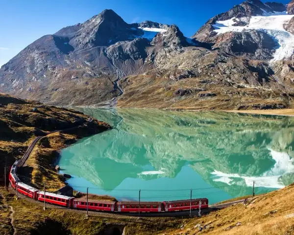 Czerwony pociąg wije się wzdłuż turkusowego jeziora u podnóża alpejskich szczytów. Idealne miejsce na trekking w alpach.