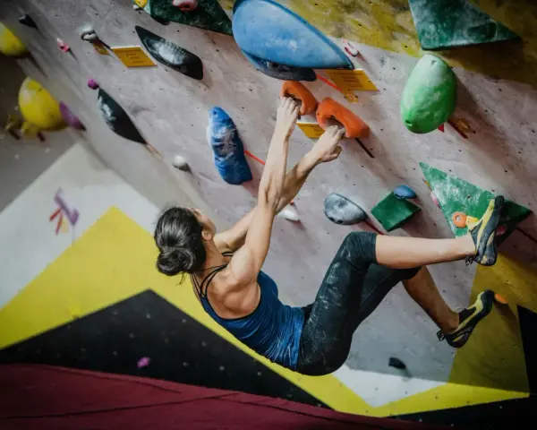 Kobieta wspina się po kolorowej ścianie, szukając chwytów. To właśnie bouldering – sportowa wspinaczka bez liny, na niskich ścianach.