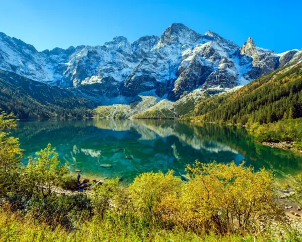 Turkusowe Morskie Oko odbija ośnieżone szczyty Tatr. Jesienne drzewa otaczają taflę wody.