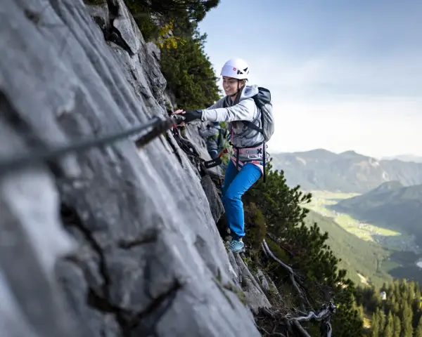 Kobieta w kasku i uprzęży pokonuje skalną ścianę, korzystając z liny. To właśnie jest via ferrata – bezpieczna droga wspinaczkowa.