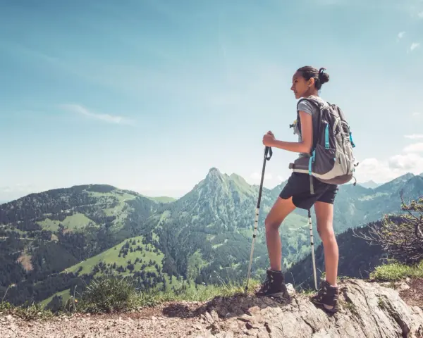 Młoda kobieta z plecakiem i kijkami trekkingowymi podziwia górski krajobraz. Pokazuje, jak chodzić z kijkami trekkingowymi, by czerpać radość z wędrówki.