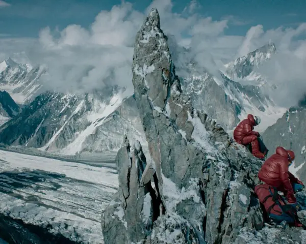 Dwóch alpinistów w czerwonych kurtkach odpoczywa na skalistym szczycie, podziwiając lodowcową dolinę i ośnieżone góry.