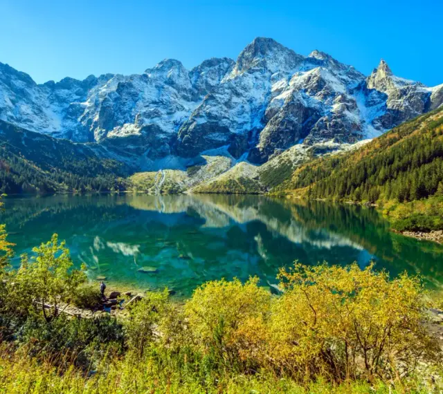 Turkusowe Morskie Oko odbija ośnieżone szczyty Tatr. Jesienne drzewa otaczają taflę wody.