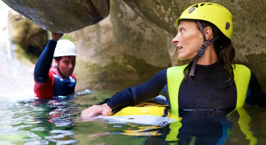 Kobieta w żółtym kasku i piankowym stroju zanurzona w wodzie, obok mężczyzna w białym kasku. To właśnie canyoning – przygoda w kanionie.