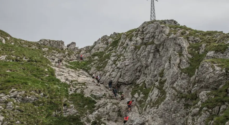 Grupa turystów wspina się po skalistym zboczu w kierunku krzyża na szczycie. Trudno określić, ile idzie się na Giewont, ale widok jest imponujący.