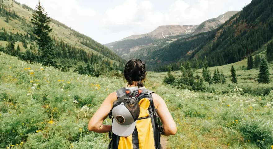 Kobieta z plecakiem wyrusza na trekking przez kwiecistą dolinę, podziwiając górskie krajobrazy.