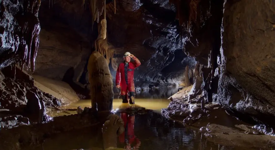 Eksplorator w jaskini, zanurzony po kolana w wodzie, odkrywa tajemnice speleologii. Stalagmity i nacieki tworzą niezwykły krajobraz.