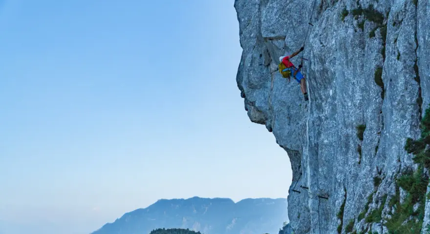 Wspinacz pokonuje via ferrata niemcy, zmagając się z pionową ścianą skalną. W tle piękne góry.